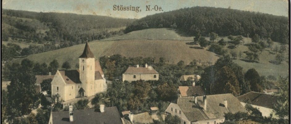 Eine Luftaufnahme des Dorfes Stossing mit einer Kirche, Häusern und einer grünen Landschaft mit Bäumen und Hügeln im Hintergrund.