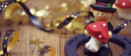 Eine Miniaturfigur eines lächelnden Jungen, der einen Hut trägt und einen roten Pilz in der Hand hält, steht auf einer Holzoberfläche. Ein Hufeisen befindet sich in der Nähe der Figur. Auf der Holzoberfläche befinden sich goldene Glitzer und Sterne.