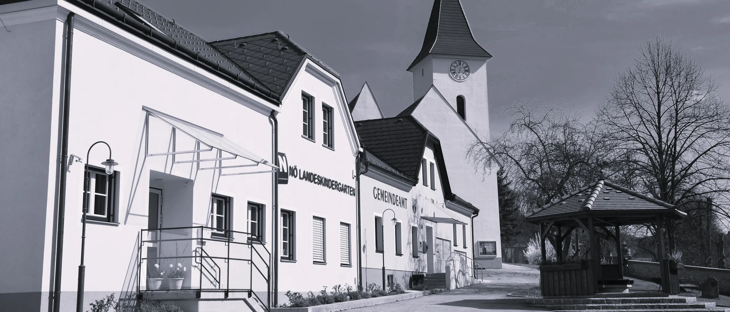 Ein Schwarz-Weiß-Bild einer Kirche mit einem Kirchturm und einer Uhr. Die Kirche ist von einem großen Gebäude umgeben, auf dem Schild steht 'No Landeskindergarten Gemeindeamt'.