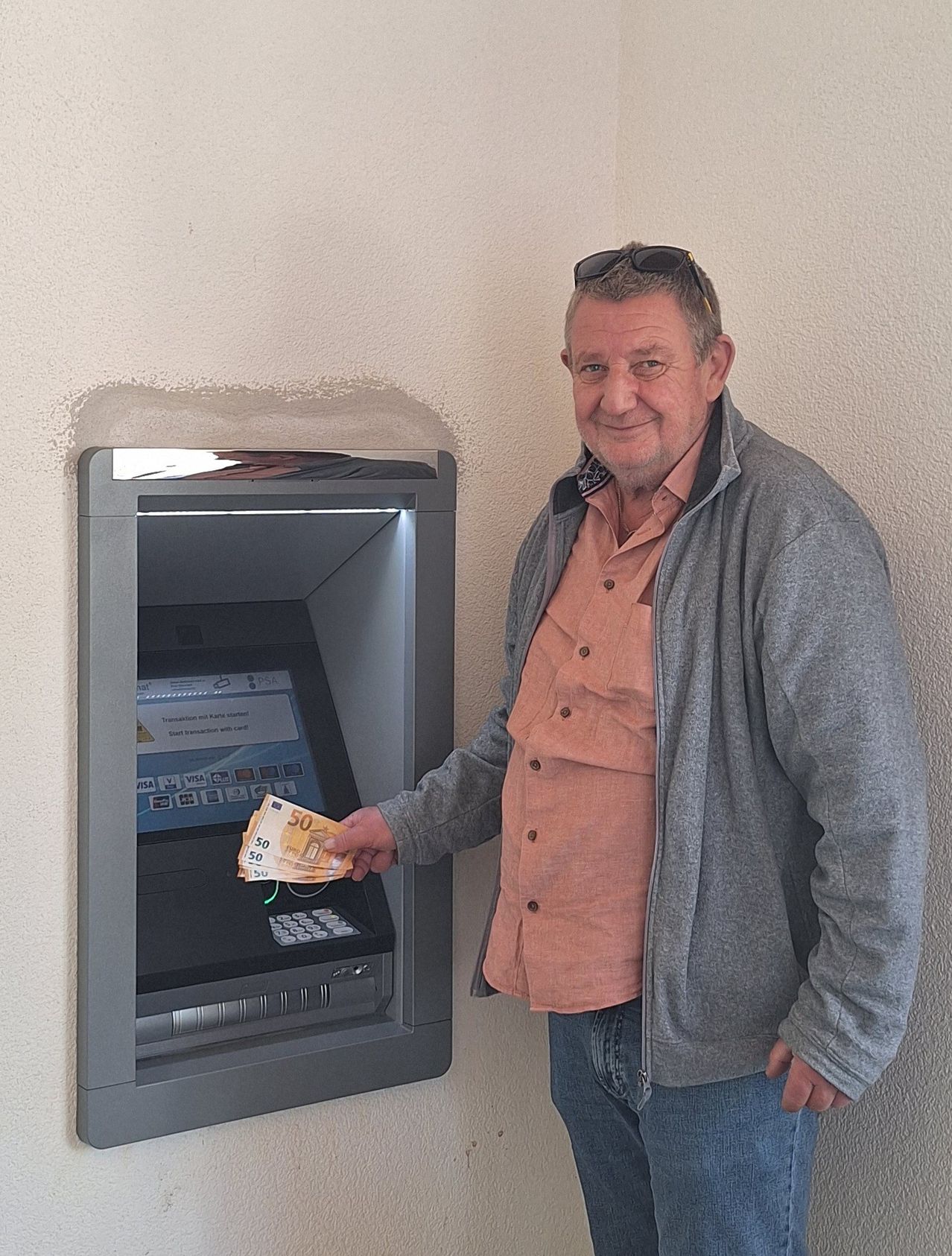 Ein Mann mit Sonnenbrille auf dem Kopf steht neben einem Geldautomaten und lächelt. Er hält Geld in der Hand.
