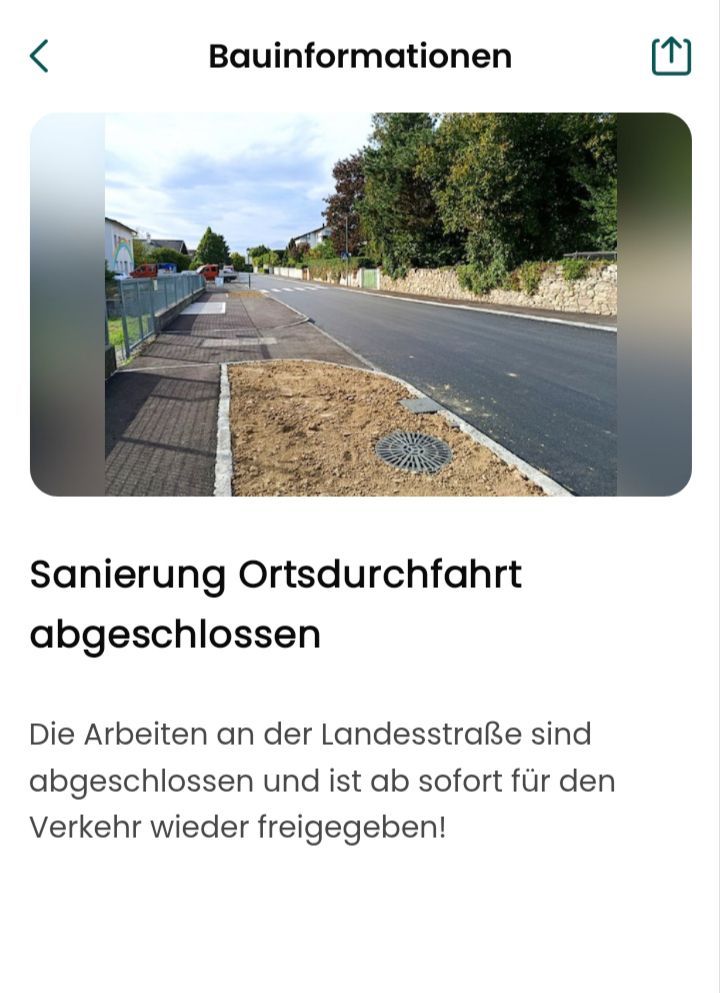 Eine neu gepflasterte Straße mit einem Bürgersteig und einem Abflussrost. Das Gebiet ist von einer Steinmauer und Grünflächen umgeben. Der Textüberlagerung lautet 'Sanierung Ortsdurchfahrt abgeschlossen'.