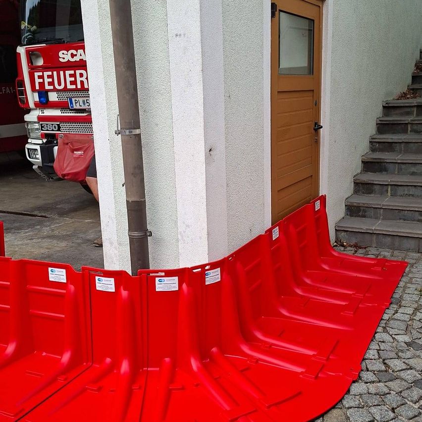 Ein roter Feuerwehrwagen steht in einer Garage, mit einer roten Barriere auf dem Boden vor einem Gebäude.