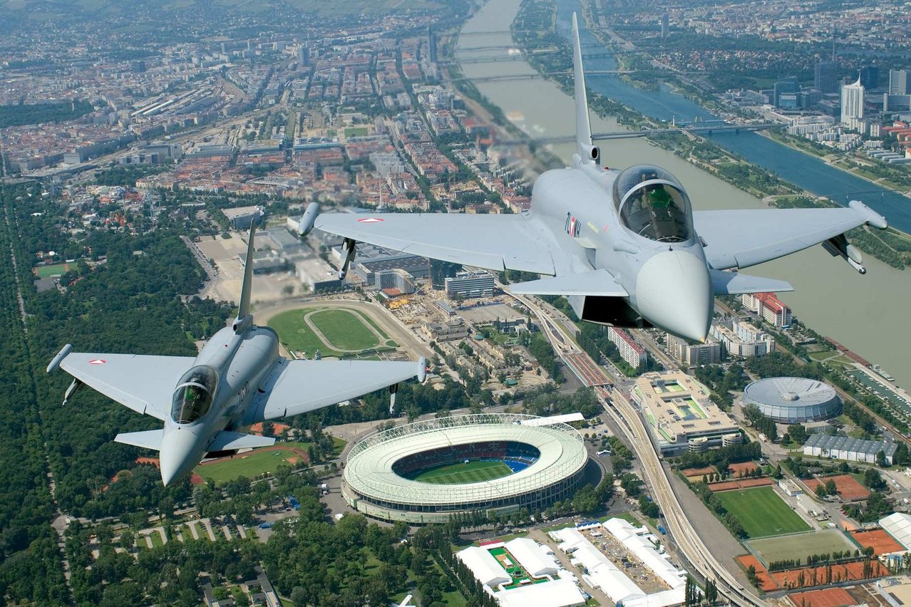 Zwei Kampfjets fliegen über eine Stadt. Ein Jet ist über dem anderen, und beide haben ihre Landeklappen eingefahren. Die Stadt darunter hat ein Stadion, Gebäude und Bäume.