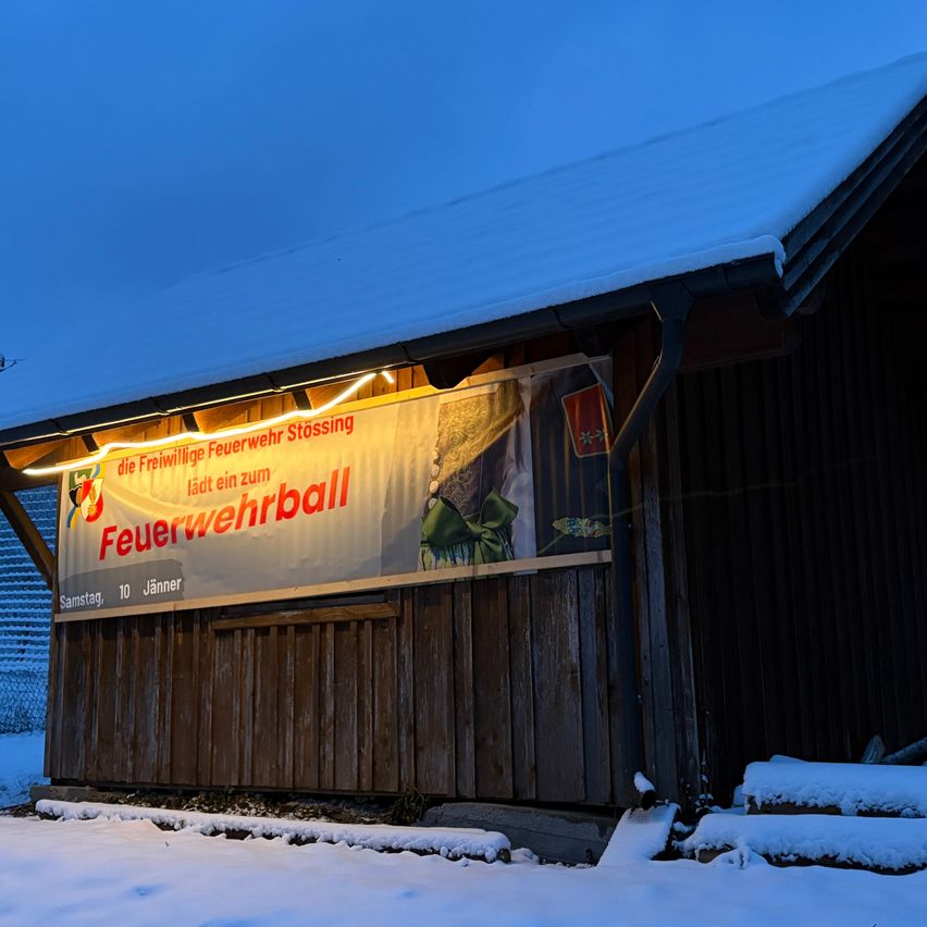 Ein Banner mit dem Text Feuerwehrball hängt an der Seite eines Holzgebäudes. Es ist bei Nacht beleuchtet.
