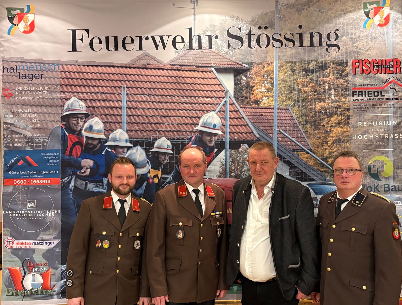 Vier Männer in Uniformen stehen vor einem Schild, das 'Feuerwehr Stossing' besagt. Die Männer posieren für ein Foto mit einem Haus und Bäumen im Hintergrund.