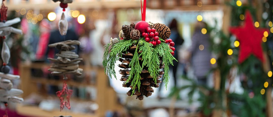 Ein Weihnachtsschmuck hängt im Vordergrund und besteht aus einem roten Ball, Tannenzapfen und roten Beeren. Es ist mit Tannennadeln geschmückt. Im Hintergrund shoppen Menschen auf einem Markt.