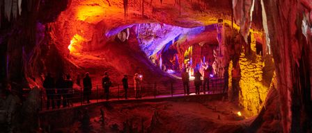 Touristen erkunden eine Höhle, die mit roten und blauen Lichtern beleuchtet ist. Die Höhle hat Stalaktiten und Stalagmiten. Einige Leute machen Fotos.