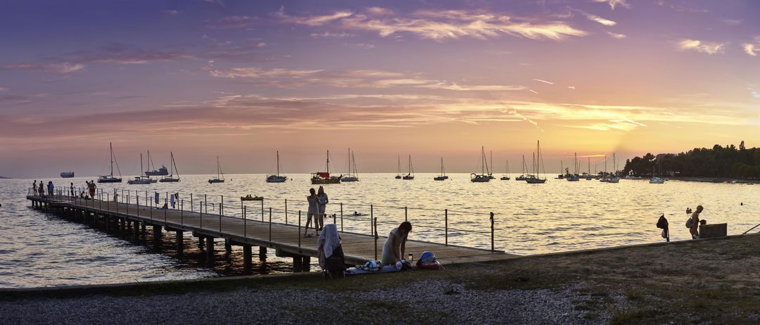Eine Gruppe von Menschen steht bei Sonnenuntergang auf einem Steg, mit Segelbooten und Booten in der Ferne. Eine Person beugt sich vor, eine andere schwimmt und zwei stehen.