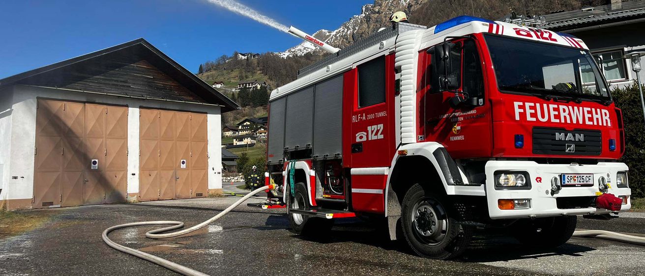 Ein roter Feuerwehrwagen mit einem Feuerwehrmann auf dem Dach spritzt Wasser aus der Düse, vor einem Gebäude mit Garage und Bergen im Hintergrund.