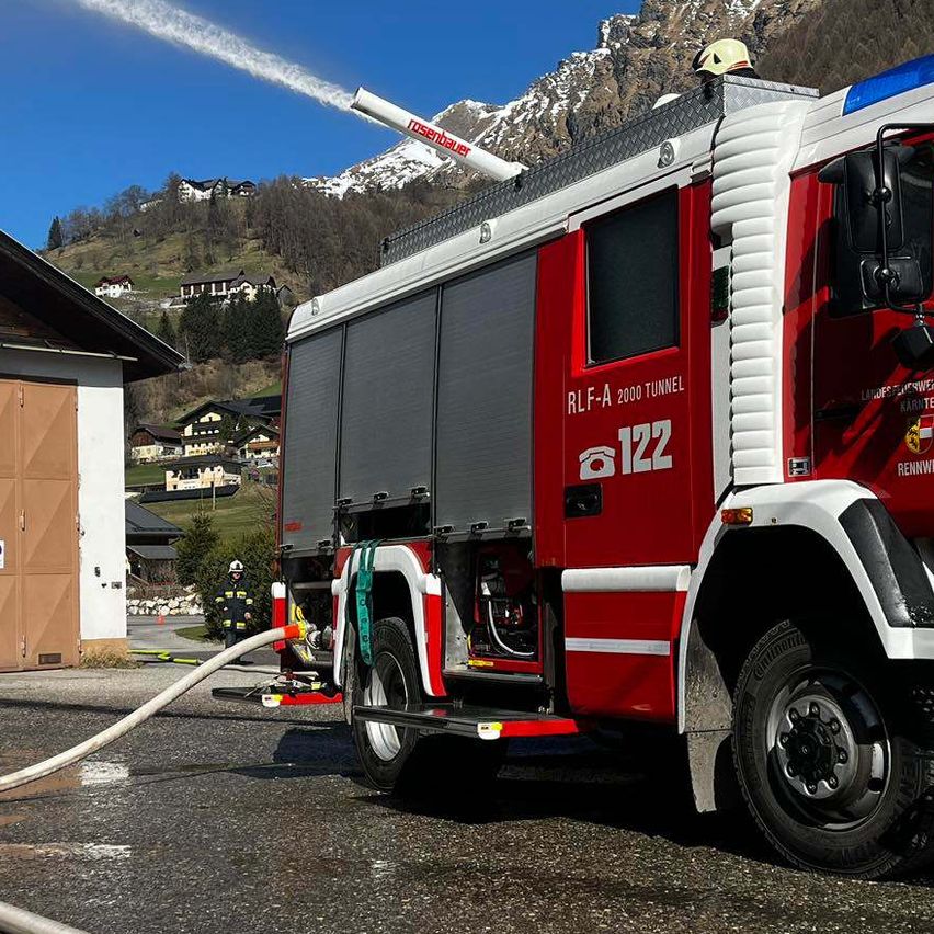 Ein roter Feuerwehrwagen mit einem Feuerwehrmann auf dem Dach spritzt Wasser aus der Düse, vor einem Gebäude mit Garage und Bergen im Hintergrund.