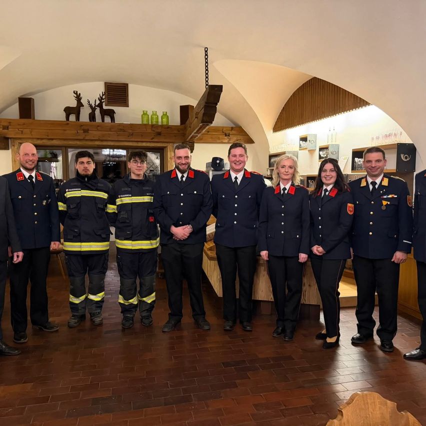 Eine Gruppe von Feuerwehrleuten in Uniform steht gemeinsam in einem Raum mit gefliestem Boden und Holzwänden und lächelt für ein Foto.