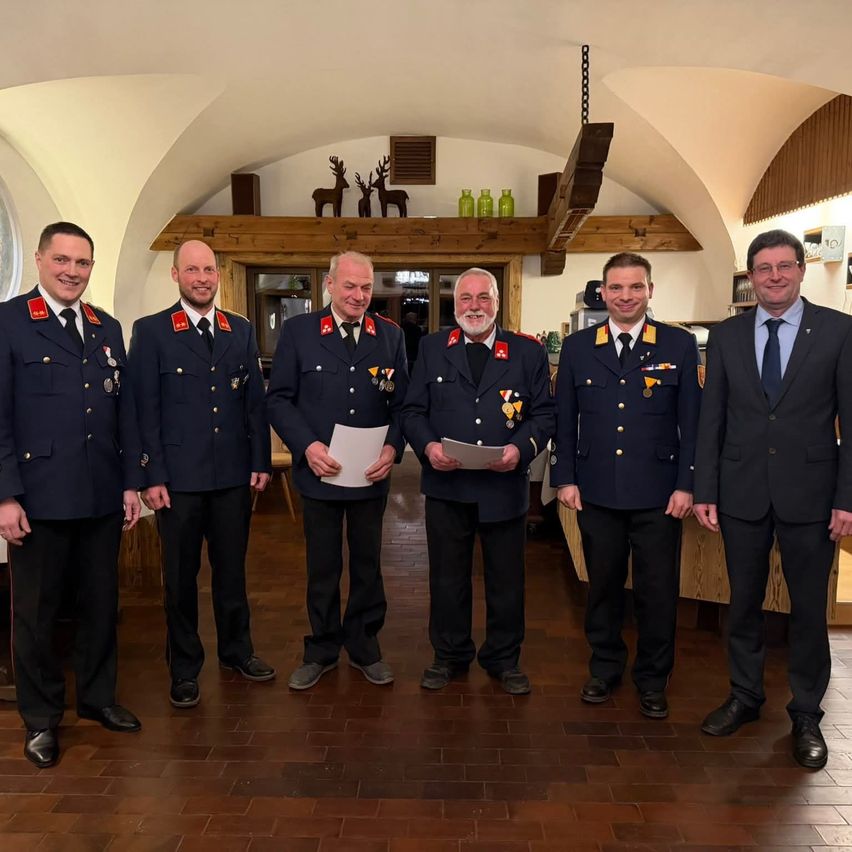 Fünf Männer in Uniformen stehen in einem Raum und lächeln für ein Foto. Zwei halten Papiere. Der Raum hat Holzböden und Balken, mit Dekorationen wie Hirschköpfen und grünen Flaschen.