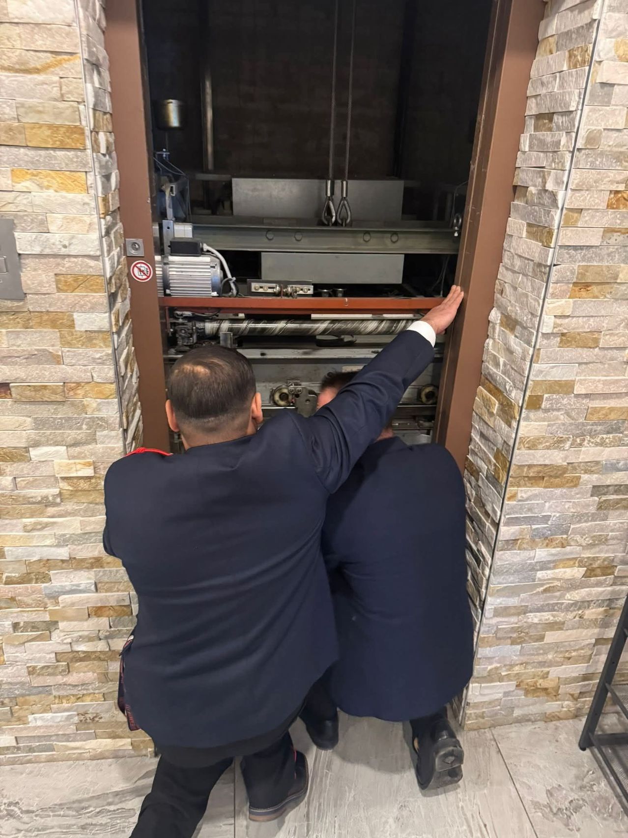 Zwei Männer in Anzügen arbeiten an der Maschinerie eines Aufzugs, einer zeigt auf die Maschinerie. Der Aufzug befindet sich zwischen Ziegelwänden.