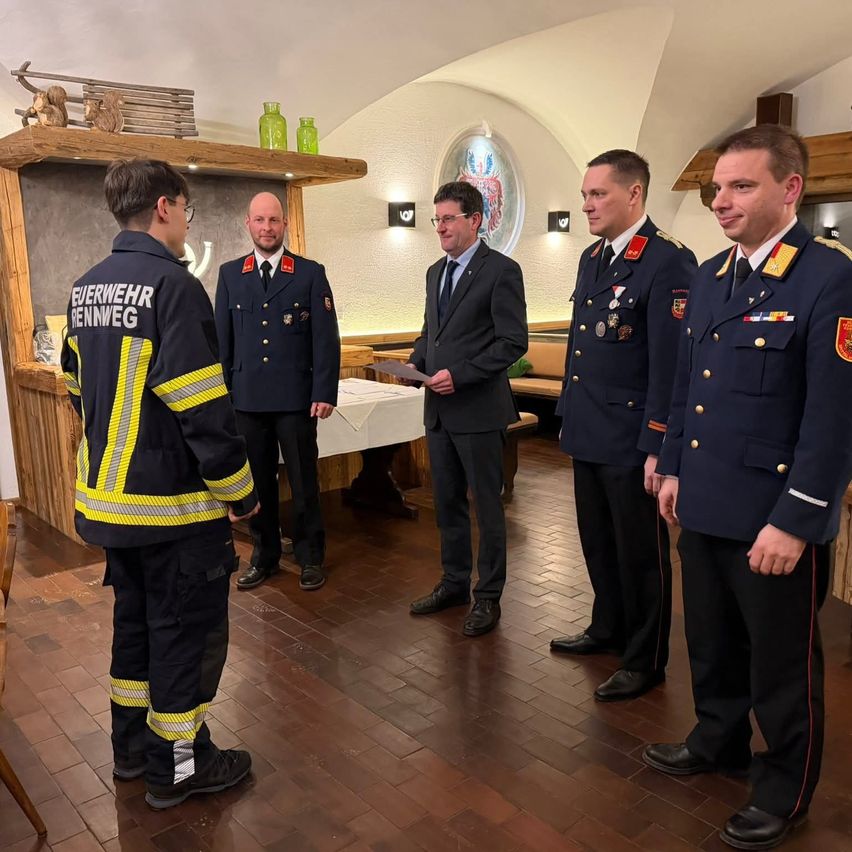 Fünf Männer in Uniformen stehen in einem Raum. Drei tragen Militäruniformen, einer ist Feuerwehrmann und einer trägt einen Anzug. Sie scheinen sich zu unterhalten.