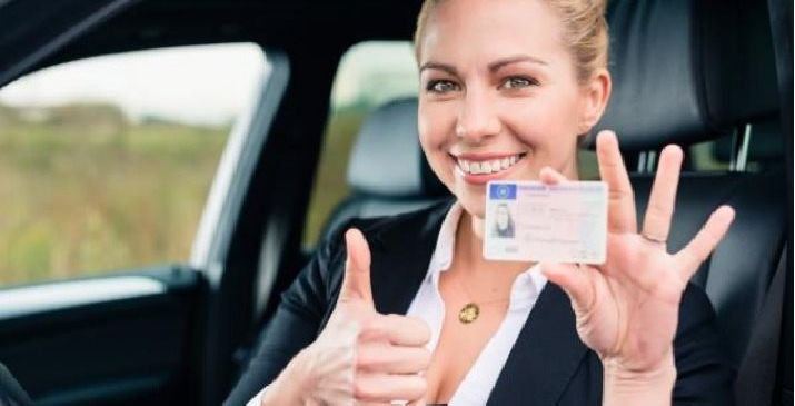 Eine lächelnde Frau in einem Auto zeigt mit dem Daumen nach oben und hält eine Karte, möglicherweise einen Führerschein, in der linken Hand.