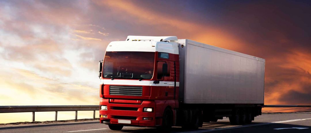 Ein großer roter und weißer Lkw fährt mit einem Sonnenuntergang im Hintergrund die Straße entlang. Der Himmel ist blau mit orangefarbenen und rosa Farbtönen.