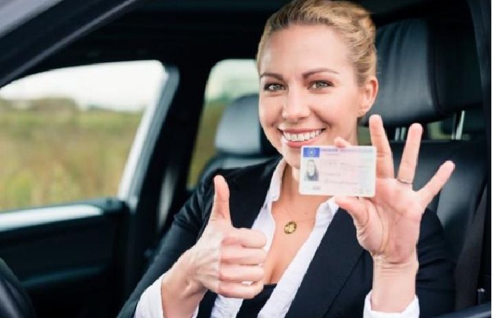 Eine Frau sitzt in einem Auto, lächelt und zeigt ihre Daumen hoch, während sie einen Reisepass in der Hand hält.