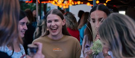 Eine Gruppe von Menschen versammelt sich unter einem Zelt. Eine Frau in einem braunen Shirt lächelt, während eine andere Frau isst. Eine Person hält eine Kamera.