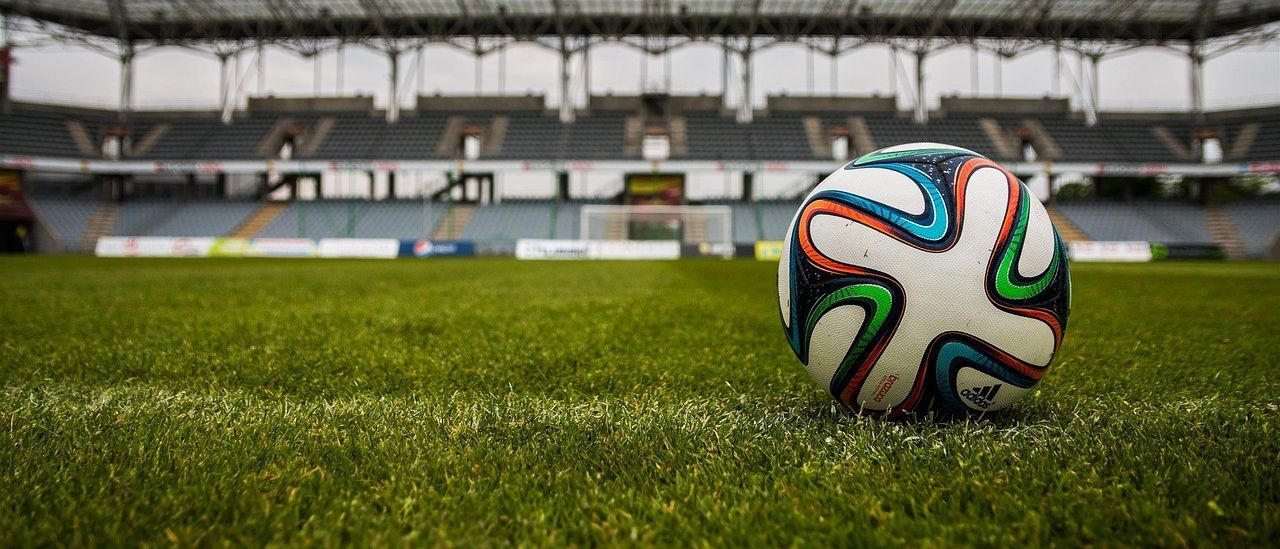 Ein Fußball liegt auf dem grünen Rasen vor einem leeren Stadion mit Metalldach.