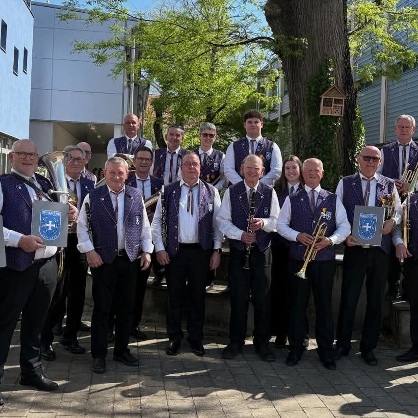 Eine Brassband in lila Westen posiert für ein Foto vor einem Gebäude mit Bäumen und einem Vogelhäuschen.