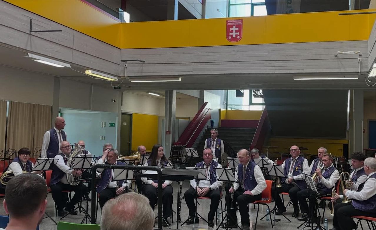 Eine Brassband tritt in einer Halle auf. Die Musiker sitzen und halten verschiedene Instrumente. Das Publikum sitzt vor ihnen. Die Halle hat eine Treppe und ein gelbes Geländer.
