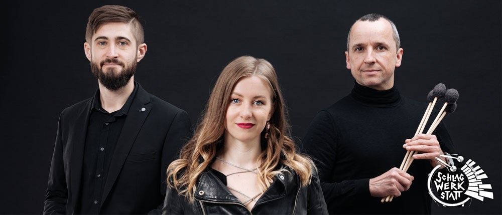 Drei Musiker stehen vor einem schwarzen Hintergrund, ein Mann mit Bart, eine Frau in einer Lederjacke und ein Mann mit Drumsticks.