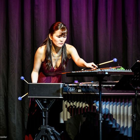 Eine Frau in einem burgunderroten Kleid spielt auf der Bühne ein Xylophon mit einem lila Vorhang im Hintergrund.