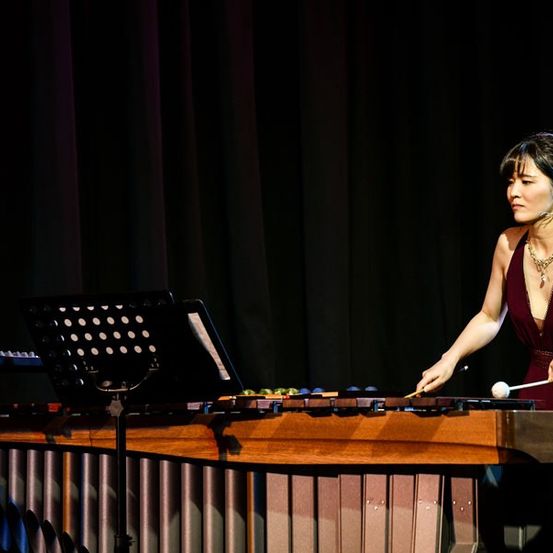 Eine Frau spielt auf der Bühne eine Marimba und trägt eine Halskette und Ohrringe. Hinter ihr steht ein Klavier und ein schwarzer Vorhang.