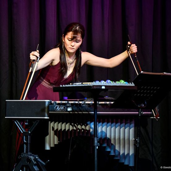 Eine Frau spielt auf der Bühne Marimba und Xylophon, trägt ein weinrotes Kleid und eine Halskette und hält Schlägel.