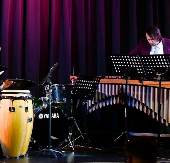 Zwei Musiker treten auf der Bühne auf, einer spielt Xylophon und der andere Schlagzeug. Die Frau sitzt und spielt die Trommeln, während der Mann hinter dem Xylophon steht.