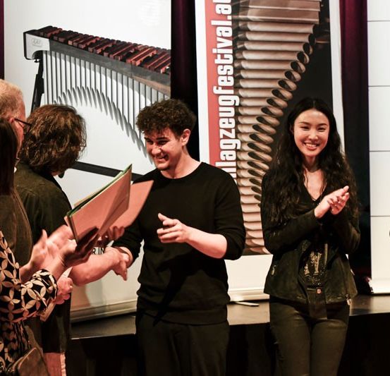 Eine Gruppe von Menschen steht auf der Bühne mit Musikinstrumenten im Hintergrund. Ein Mann und eine Frau klatschen in die Hände.