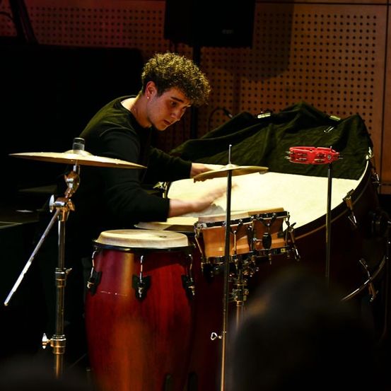 Ein junger Mann spielt in einem schwach beleuchteten Raum Trommeln. Er schlägt verschiedene Trommeln und Becken. Hinter ihm hängt ein schwarzer Vorhang an einer Holzwand.