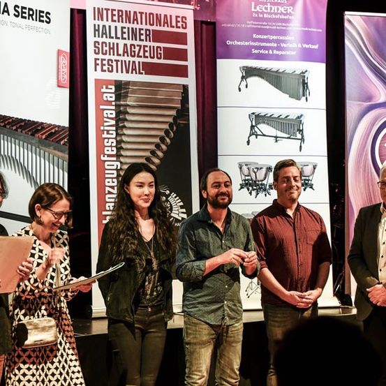 Fünf Personen stehen auf der Bühne und halten Urkunden in den Händen, begleitet von einem Banner mit der Aufschrift 'Internationales Halleiner Schlagzeug Festival'. Sie scheinen Auszeichnungen zu erhalten.