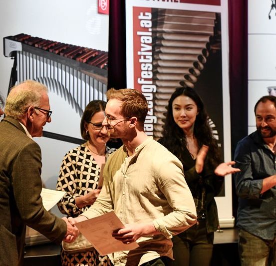Eine Gruppe von Menschen steht auf der Bühne. Ein Mann überreicht einem anderen einen Zettel, während andere klatschen. Hinter ihnen sind Poster mit Musikinstrumenten.