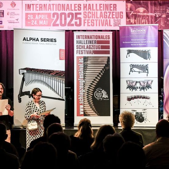 Zwei Personen auf der Bühne beim Internationalen Halleiner Schlagzeug Festival 2025. Eine Frau hält ein Buch, und ein Mann ein Papier. Hinter ihnen sind Banner mit Informationen über das Festival.