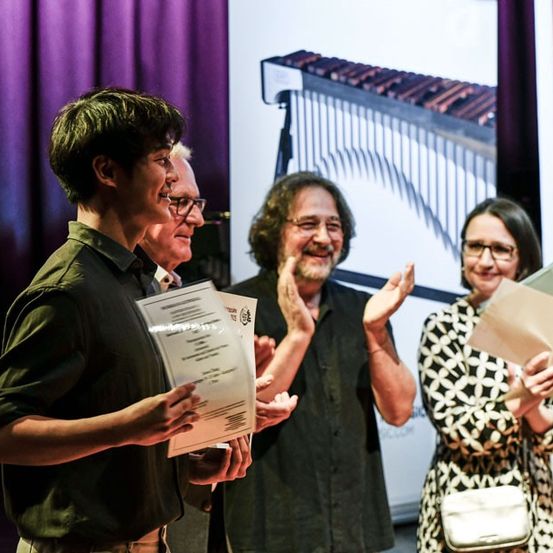 Vier Personen stehen auf der Bühne, möglicherweise bei einer Preisverleihung. Sie halten Papiere und klatschen, mit einem großen Xylophon im Hintergrund.
