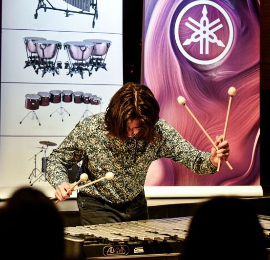 Ein Musiker spielt auf der Bühne ein Xylophon, mit einem Yamaha-Logo im Hintergrund. Das Publikum sitzt vor der Bühne und beobachtet die Aufführung.