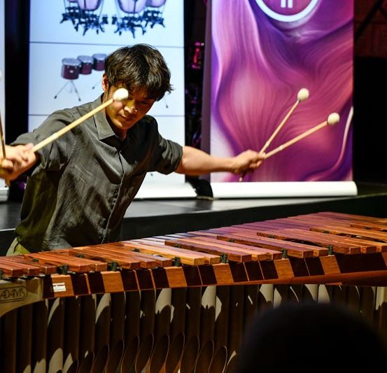 Ein Mann auf der Bühne spielt ein hölzerne Xylophon mit zwei Schlägeln und tritt vor einem Publikum auf.