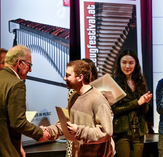 Eine Preisverleihung mit einem Mann in Anzug, der einem Mann in Pullover eine Auszeichnung überreicht. Hinter ihnen klatschen zwei Frauen. Im Hintergrund ein Banner mit dem Text 'Guggfestival' und Musikinstrumente.