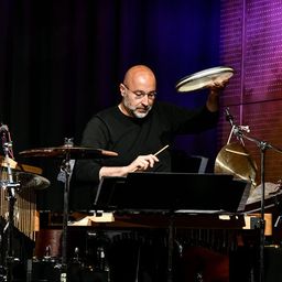 Ein kahlköpfiger Mann mit Brille und schwarzem Shirt spielt ein Marimba auf der Bühne. Er hält einen Drumstick und hat andere Musikinstrumente vor sich.