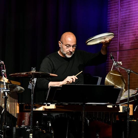 Ein kahlköpfiger Mann mit Brille und schwarzem Shirt spielt ein Marimba auf der Bühne. Er hält einen Drumstick und hat andere Musikinstrumente vor sich.