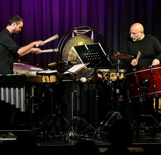 Zwei Männer spielen auf der Bühne verschiedene Musikinstrumente. Ein Mann spielt Schlagzeug, der andere eine große Gong.
