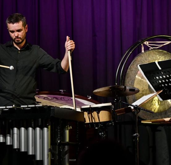 Ein Mann spielt Xylophon und Gongs auf einer Bühne mit einem lila Hintergrund. Er trägt ein schwarzes Hemd und hält Trommelstöcke.