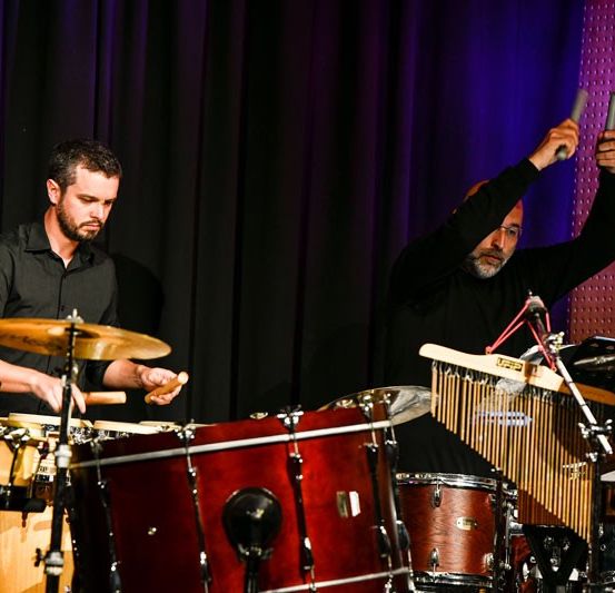Zwei Männer spielen auf der Bühne mit Trommeln und Perkussionsinstrumenten. Einer hält Stöcke und der andere spielt eine Marimba. Sie stehen vor einem schwarzen Vorhang.