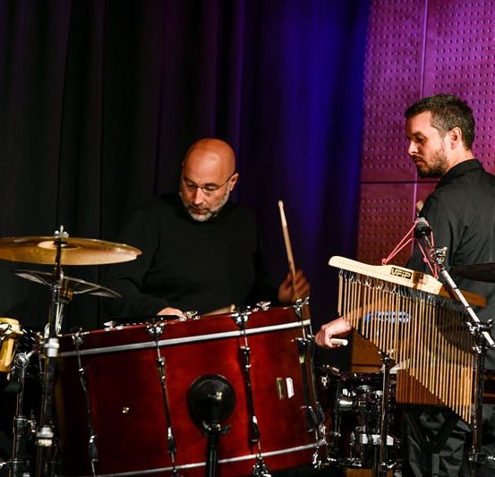 Zwei Musiker spielen auf der Bühne mit Trommeln und Xylophonen unter Scheinwerfern. Ein Mann ist kahl und hält Trommelstöcke, der andere hat einen Bart und spielt Xylophon.
