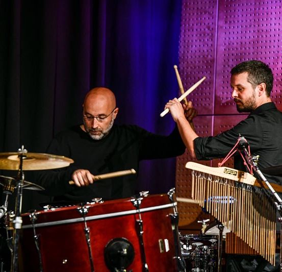 Zwei Männer spielen auf der Bühne mit Trommeln und einem Xylophon, einer spielt die Trommel, der andere das Xylophon, die Bühne ist mit lila Lichtern beleuchtet.