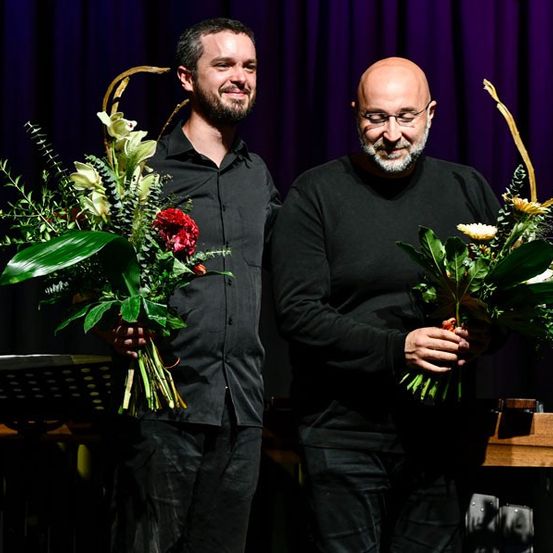 Zwei Männer auf der Bühne, beide halten Blumensträuße in den Händen und lächeln in die Kamera. Einer hat einen Bart, der andere trägt eine Brille. Hinter ihnen ein lila Vorhang und ein Holzpult für Noten.