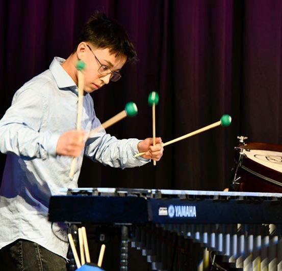 Ein junger Mann mit Brille und blauem Hemd spielt Xylophon und Schlagzeug auf einer Bühne.
