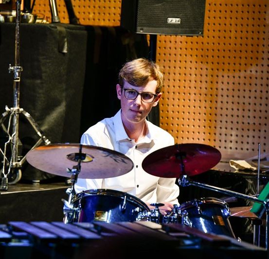 Ein junger Mann spielt Schlagzeug auf einer Bühne mit einem Lautsprecher und Becken hinter ihm. Er trägt eine Brille und ein weißes Hemd.