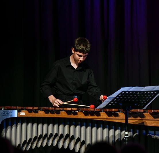 Ein junger Mann spielt Xylophon und hält zwei rote Schlägel in den Händen. Vor ihm steht ein Notenständer. Die Bühne ist mit einem dunklen Hintergrund ausgestattet und er wird von Bühnenlichtern beleuchtet.